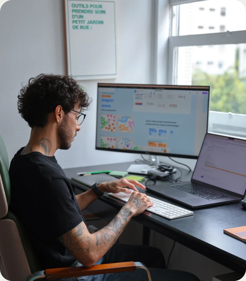Guilherme at his desk, typing, with a monitor and a laptop open in front of him.