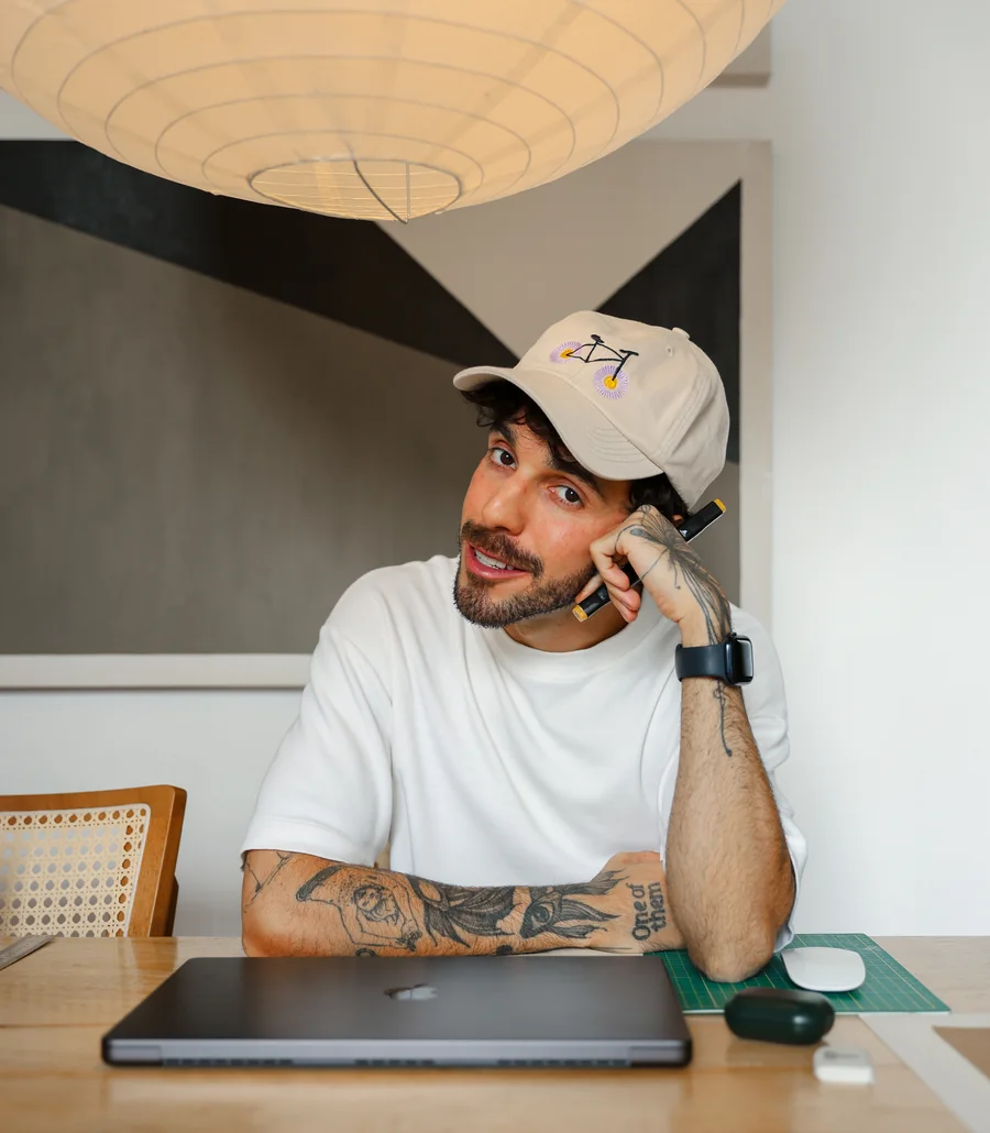 Guilherme in a white cap, resting his chin on his hand at a table with a closed laptop.